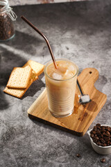 A glass with ice cubes and milk coffee on a wooden board, with brass straw
