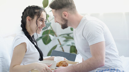 happy man bringing breakfast to smiling girlfriend with braids in bed.