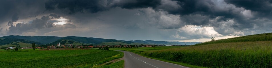 Fototapeta na wymiar Panoramic image, asphalt road leading to a small hungarian village in Transylvania, Romania, dramatic gathering storm clouds in the background.