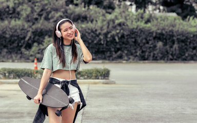 Beautiful sportive hipster happy Asian woman wearing casual shirt and shorts with headphone to listen music while holding skateboard as hobby in free time day on holiday, smiling at outdoor park. © Ann Rodchua