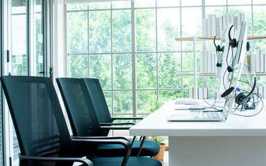 Background of interior modern office consist of table and book shelves beside window with tree and nature outside, computers and ergonomic chairs for call center service with nobody in room.