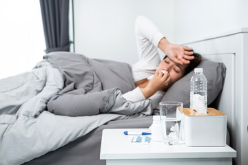 Asian woman feeling sick and sleeping in blanket on the bed while using tissue to cough, sneezing