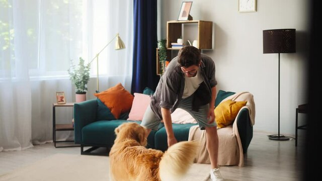 Man training his dog, throwing small ball to obedient golden retriever in living-room. Energetic puppy running after toy. Playing and spending time together with beloved pet at home during lockdown. 