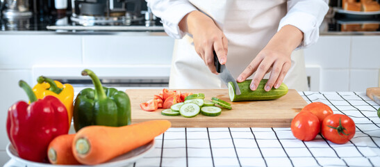 Asian housewife wearing apron and using knife to slice cucumber and tomato on chopping board