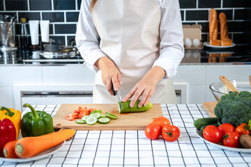 Asian housewife wearing apron and using knife to slice cucumber and tomato on chopping board