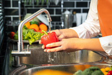Asian housewife wearing an apron and standing to washing bell pepper and vegetables with water in sink