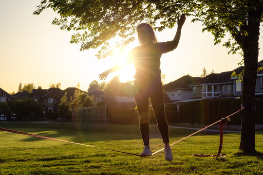 Adventurous White Caucasian Adult Woman walking on a Slackline between trees in a neighborhood park. Sunny Sunset. Surrey, Vancouver, British Columbia, Canada.