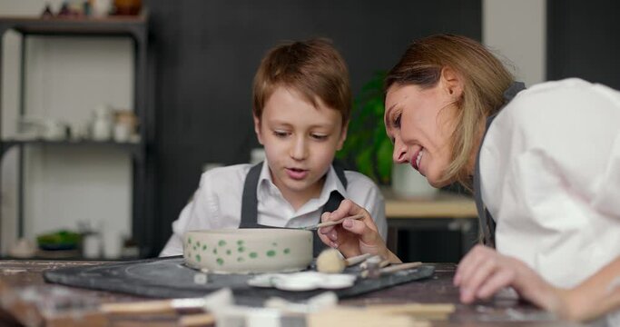 Mother assisting her son in painting bowl in studio. Pottery class workshop. Mom and child spend time together doing creative work. Slow motion