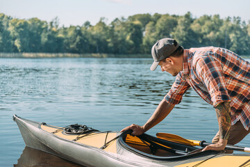 Young man with a tattoo in a cap and shirt launching a kayak. - 441212582