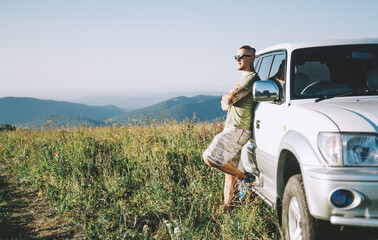 Young man in sunglasses and a tattoo on his arm stands near an off-road vehicle in nature in the mountains.