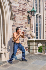 Dressing in jeans and leather sneakers and shirtless, a handsome, muscular guy is waving arms and exercising to counterattack