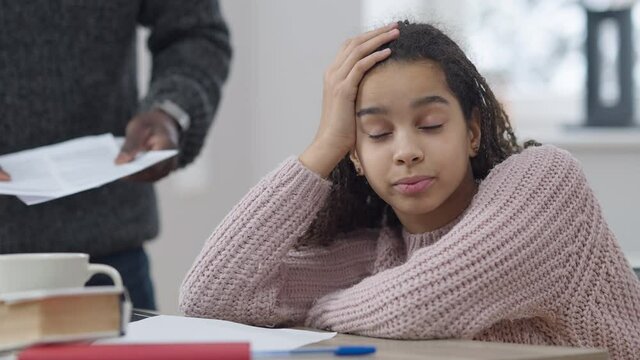 Misbehaving African American adolescent girl grimacing saying bla-bla as unrecognizable man gesturing at background. Unknown angry father scolding bored daughter at home. Adolescence and education