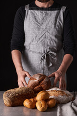 A man baker with a beard in a gray apron stands against a black background and holds, breaks, cuts off delicious, crispy bread, rolls, baguette