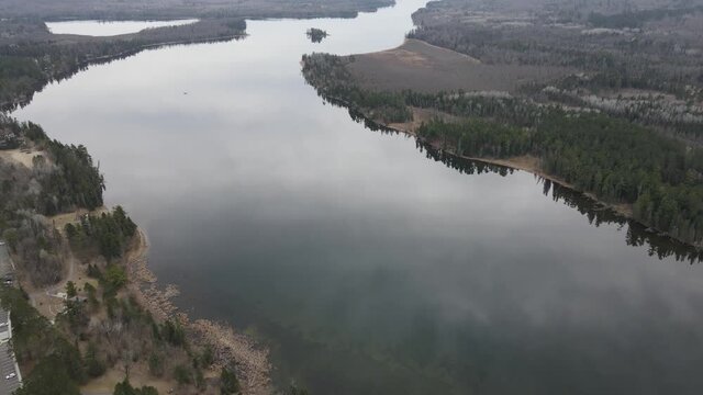 Drone clip slowly flying over Lake Itasca, MN on a warm spring day in early April, water is very still. Cloud and tree reflections can be seen in lake. Landscape is still brown from winter.