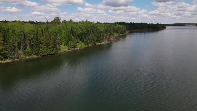 4K clip of drone circling to show beautiful, green, treed coastline next to blue rippling water at Lake Itasca, MN. Cloudy sky in background.