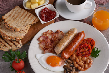 Traditional full English breakfast with fried eggs, sausages, beans, mushrooms, grilled tomatoes and bacon on a plate, Toast, butter, jam on a wooden board, Coffee and orange juice on stone background