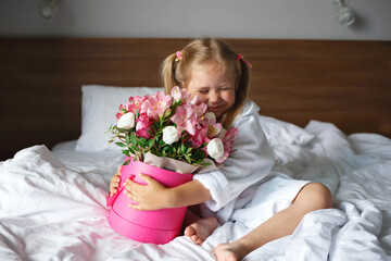 Little girl in a robe holds a box of flowers while sitting on the bed