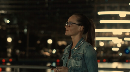 Portrait of beautiful positive young girl in casual clothes and glasses posing on the background of glowing nighttime buildings in the metropolis holding smartphone. Concept of tourist navigators apps