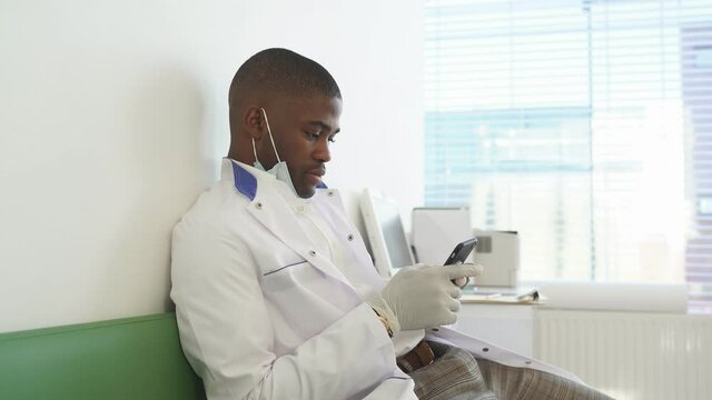 Doctor sits in medical office during free time and working on smartphone.