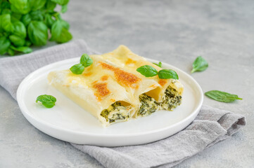 Traditional Italian cannelloni with ricotta and spinach in bechamel sauce with fresh basil leaves on a white plate on a gray concrete background. Vegetarian dish. Copy space.
