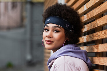 African american woman in bandana looking at camera near wooden fence outdoors