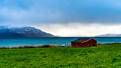 Fototapeta na wymiar Rote Hütte am Ufer des Fjord an einem stürmischen Tag in Troms, in der Nähe von Tromsö, Norwegen. interessante Lichtstimmung mit dem dunklen, aufgewühlten Meer und Wolken und Holzhaus. 