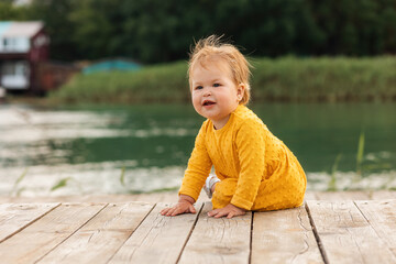 Summertime. Pretty Caucasian smiling baby girl sitting on the pier by the river. Outdoors. The...