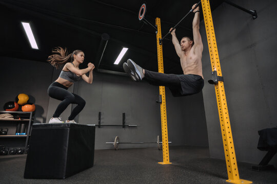 Crossfit exercise class. Young couple doing exercises at gym, having workout together. Shirtless man doing abs exercises on horizontal bar while fit woman jumping on box. Fitness and crossfit concept