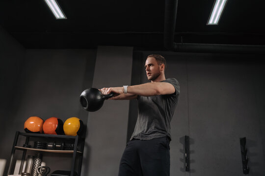 Strong muscular athlete doing kettlebell swings at crossfit gym, outfit shot on grey gym background with equipment. Weightlifter doing workout indoors, exercising. Crossfit instructor warming up