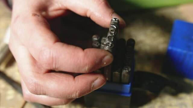 Leather craftsman at work. Close up pan of his hand choosing metal letter number stamp punch from a set