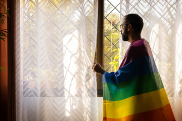 Man with lgbt flag on shoulders looking out window outdoors