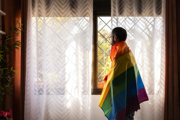 Man with lgbt flag on shoulders looking out window outdoors