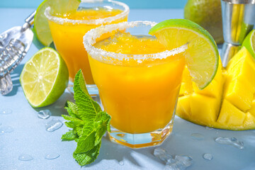 Cold summer cocktail, mango margaritas with tequila, salt lime slices, crushed ice and mint. Seasonal refreshing drink, on bright blue sun lighted background