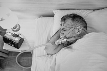 Monochrome portrait of Sleeping man with chronic breathing issues considers using CPAP machine in bed. Healthcare, Obstructive sleep apnea therapy, CPAP, snoring concept - obrazy, fototapety, plakaty