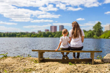 Mom and son cuddling sitting on bench by river shore at sunny summer day. Back view. Lifestyle, mothers tenderness. Son's day or mother's day concept