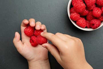 Raspberries in a saucer on a black background. The child takes raspberries from the plate and puts...