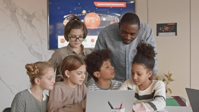 Medium slowmo of multiethnic schoolchildren and their young male Afro American teacher having fun together looking at laptop screen during Science class