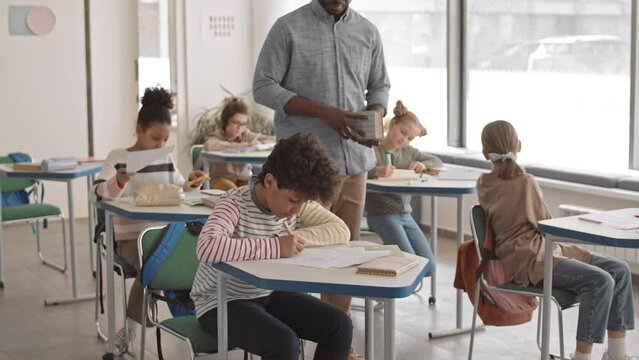 Slowmo of young male Afro American school teacher giving lesson to students in casualwear of 5 or 6 grade sitting by their desks in bright modern classroom