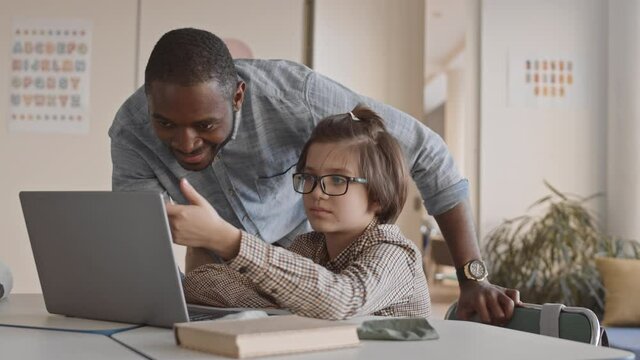 Medium slowmo of young male Afro American teacher and 5-grade schoolboy looking at laptop screen discussing homework or task