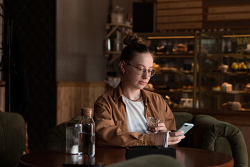 pretty young woman drinking water in cafe and wait for ordered food, using mobile phone for chatting or texting or surfing the internet