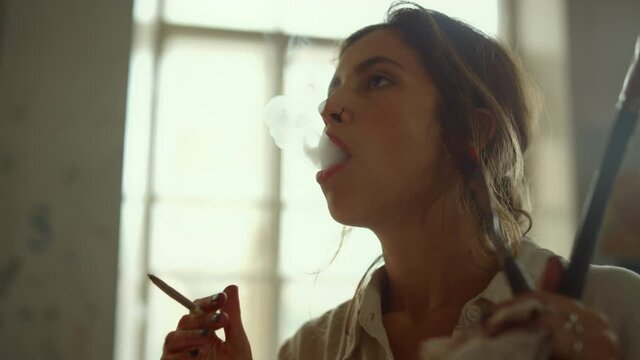Creative painter blowing smoke out in studio. Talented girl smoking indoors.