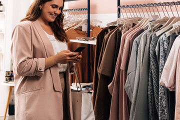 Naklejka na ściany i meble Smiling woman using cell phone while standing at a rack in a clothing store. Plus size female holding smartphone while shopping.