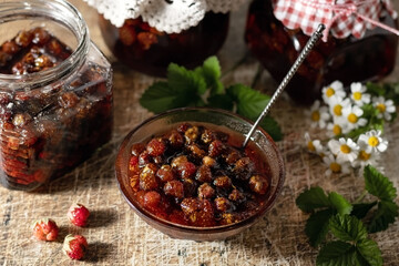 Wild strawberry jam in a glass jar with strawberries and leaves on a rustic background. Homemade strawberry marmalade and fruit