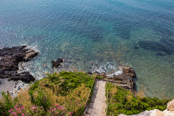 The concrete staircase surrounded by greenery descends towards the rock. Transparent sea water....