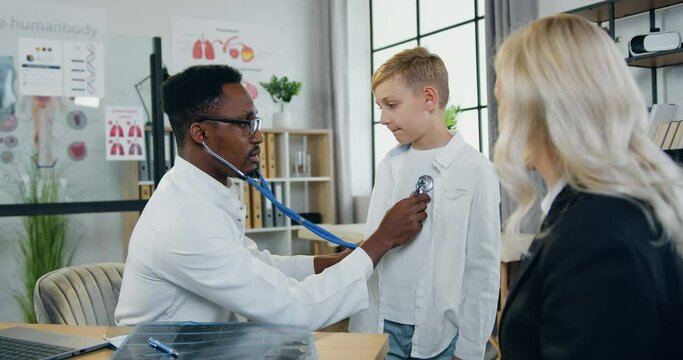 Healthcare and pediatry concept where likable confident skilled black-skinned male doctor checking boy's lungs during his scheduled visit to clinic with beautiful mother