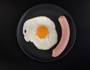 A fried egg and sausage lie on a black plate on a black background