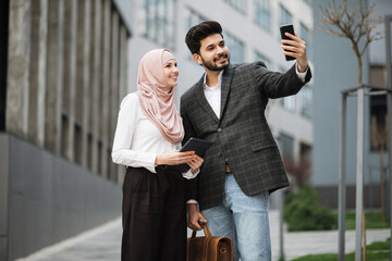Smiling arabian business colleagues standing on city street and taking selfie on modern smartphone....