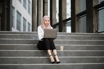 Charming woman in stylish elegant clothes and hijab sitting on stairs outdoors and using wireless laptop. Muslim female working on distance.