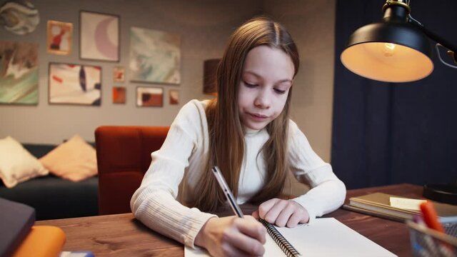 Girl kid in glasses making video call with laptop at home, writing notes, using camera zoom online virtual class, social distancing, learning remotely at school during covid coronavirus lockdown. 