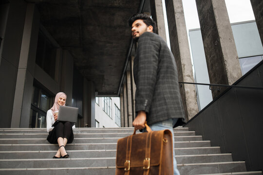 Handsome Muslim Man In Formal Wear Coming Up On Stairs While Woman In Hijab Sitting Near And Working On Laptop. Two Coworkers Doing Remote Work Outdoors.